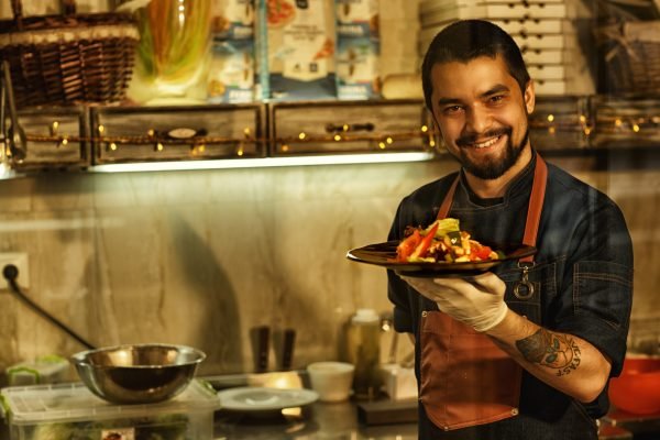 Chef smiling and showing delicious salad made of fresh vegetables. Man holding dish with salad in his hand. Background of professional restaurant kitchen with special kitchenware.